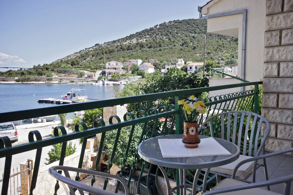 A balcony with a table and chairs, overlooking a body of water and a hillside with buildings.