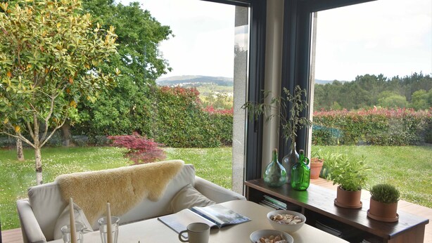 Uma área de jantar com uma mesa posta para dois, uma janela com vista para um jardim e uma estante com livros e objetos decorativos.