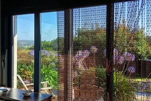 A room with a large window overlooking a garden with purple flowers and a view of the countryside.