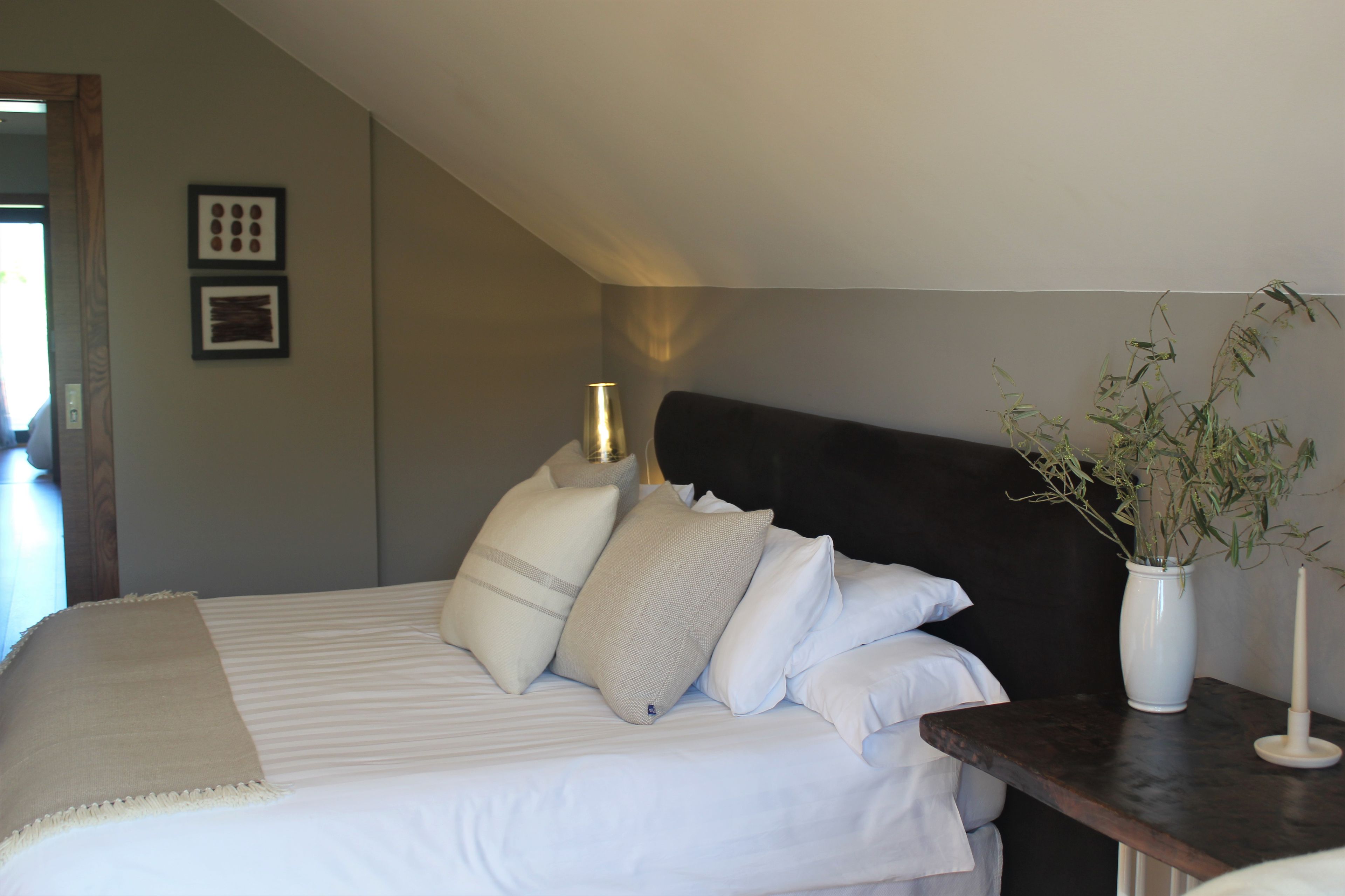 Een slaapkamer met een bed, kussens, een nachtkastje met een vaas en kaars, en een wandlamp.