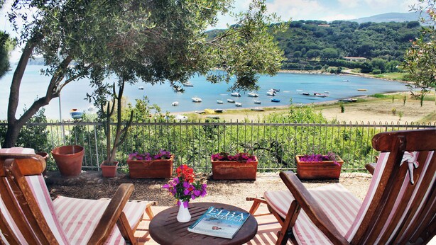Eine Terrasse mit zwei Holzstühlen, einem kleinen Tisch mit Blumenvase und einer Zeitschrift. Im Hintergrund zeigt sich ein See mit Booten und ein bewaldeter Hügel.