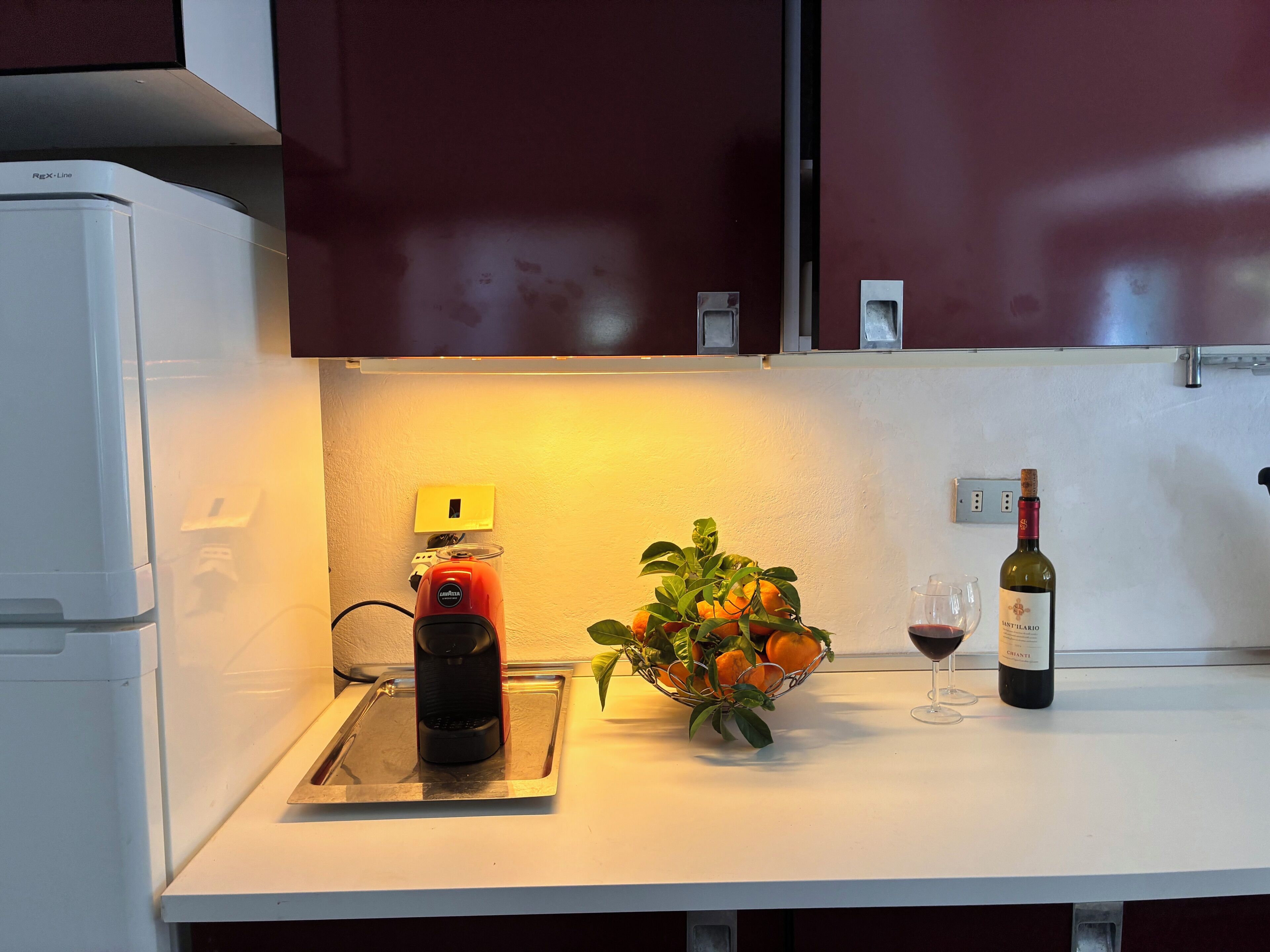 Un piano di lavoro in cucina con una macchina per il caffè, un cesto di frutta, un bicchiere da vino e una bottiglia di vino.