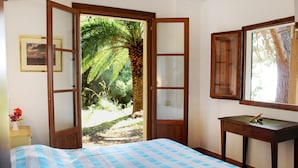 Ein Schlafzimmer mit einem Bett, einem Nachttisch aus Holz, einem Schreibtisch und einem großen Fenster mit Blick auf Palmen.