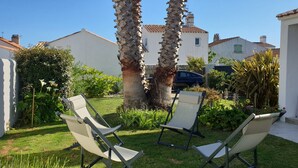 Um jardim com duas cadeiras de descanso, uma palmeira e um edifício branco com telhado de azulejos.