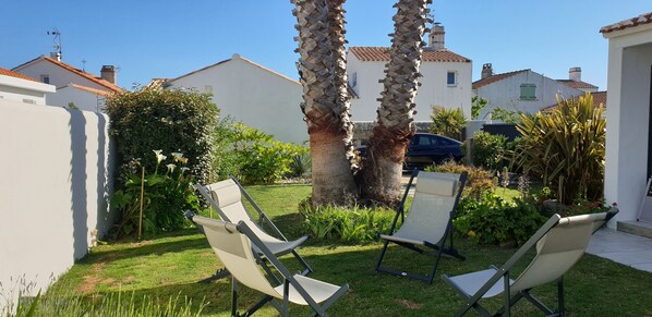 A garden with two lounge chairs, a palm tree, and a white building with a tiled roof. - house / villa - 4 rooms - 6/8 persons (Noirmoutier-En-L'île)