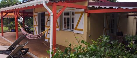 A small, single-story building with a red and white exterior, a covered patio area with a hammock and lounge chairs, and a garden in the foreground.
