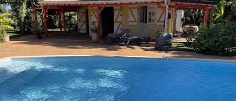 Un espace aménagé au bord de la piscine, comprenant un coin salon couvert, une piscine et une végétation luxuriante.