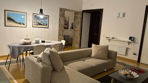 A living room with a beige sofa, a dining table with chairs, and a coffee table. There are framed pictures on the wall, a hanging light fixture, and a stone wall.