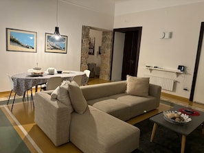 Un salon avec un canapé beige, une table à manger avec des chaises et une table basse. Les murs sont ornés de tableaux encadrés, un luminaire suspendu éclaire la pièce et un mur est recouvert de pierres.