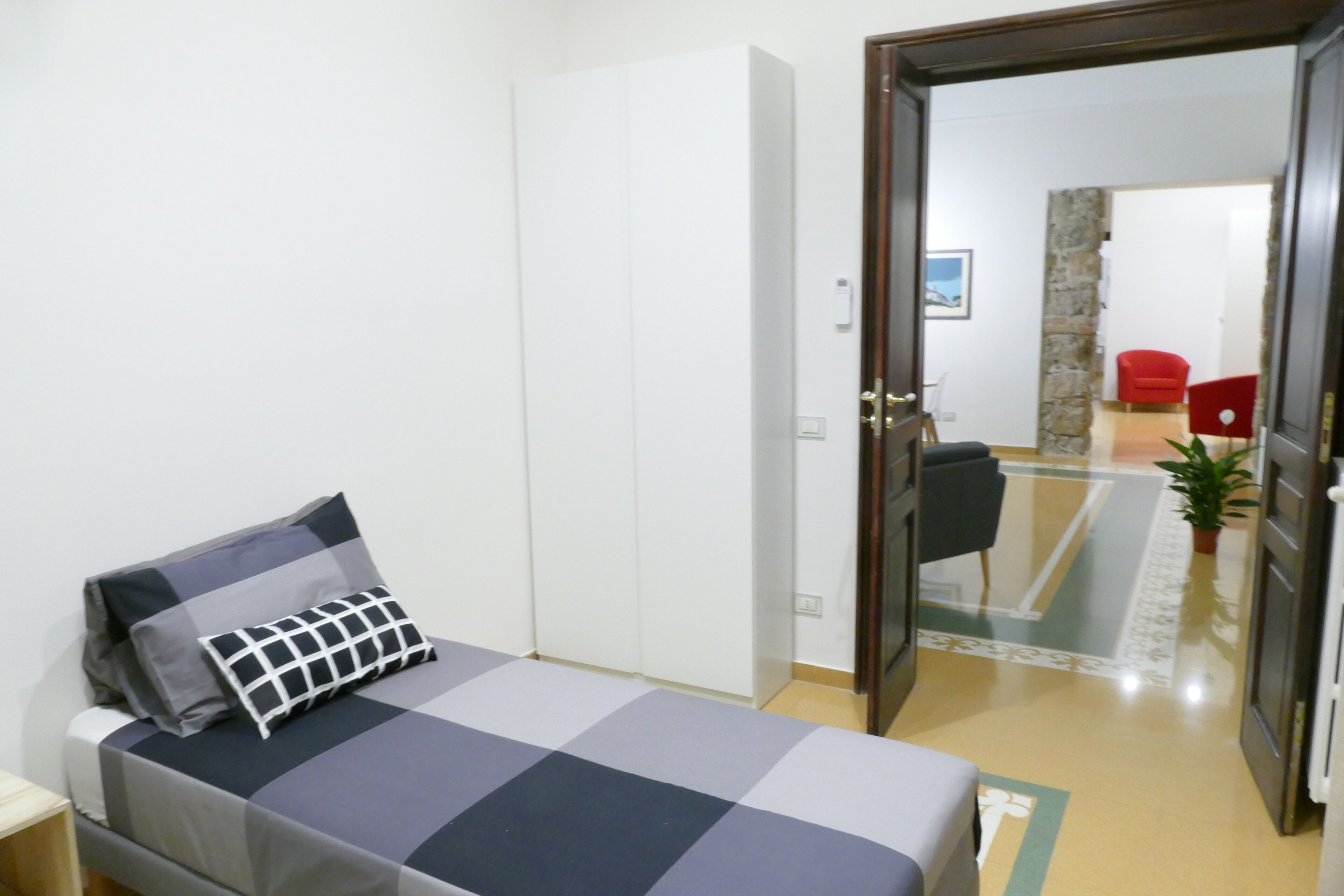 A bedroom with a bed, a wardrobe, a door leading to a living area with a red chair and a potted plant.
