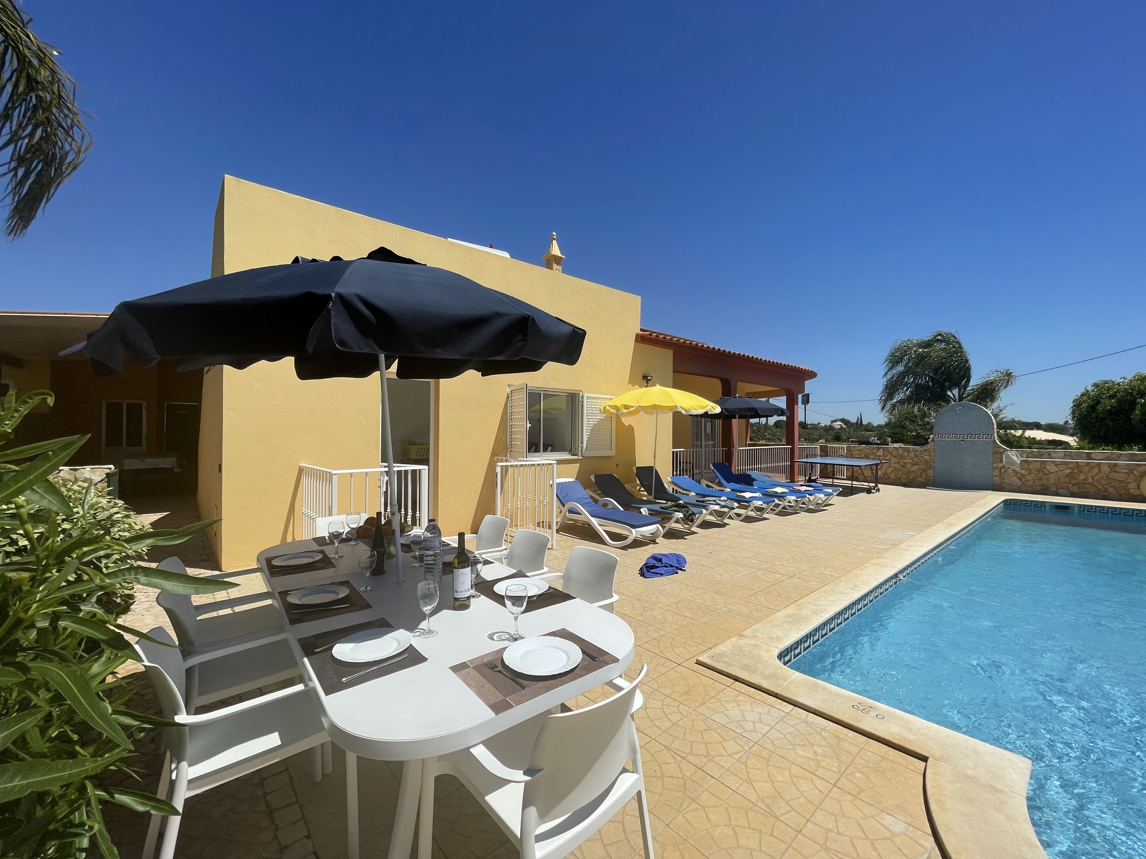 A poolside area with sun loungers, a table set for dining, and a clear blue sky.