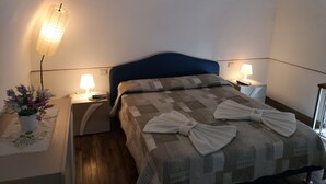 Ein ordentlich bezogenes Bett mit einer Steppdecke und zwei Handtüchern, eine Nachttischlampe, ein kleiner Tisch mit einem Blumenstrauß und eine Wandlampe.