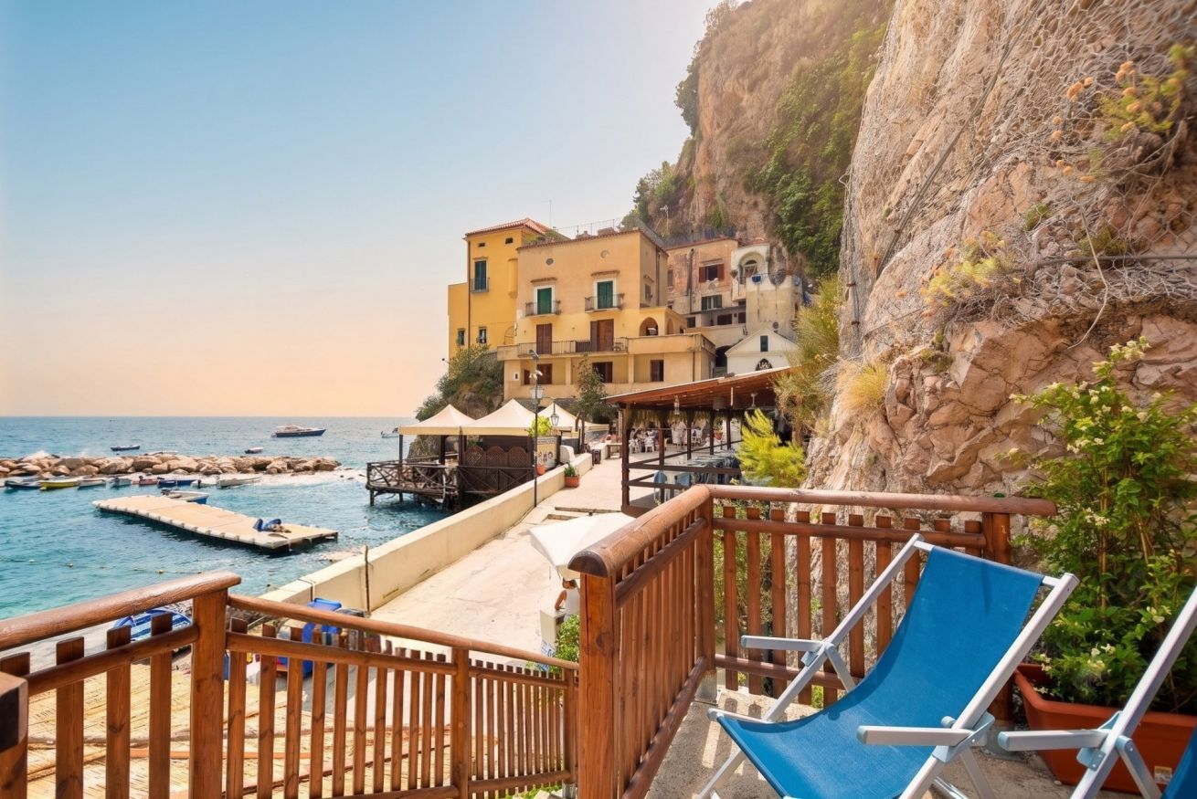 A coastal hotel with a wooden deck, blue lounge chairs, and a view of the sea and rocky cliffs.