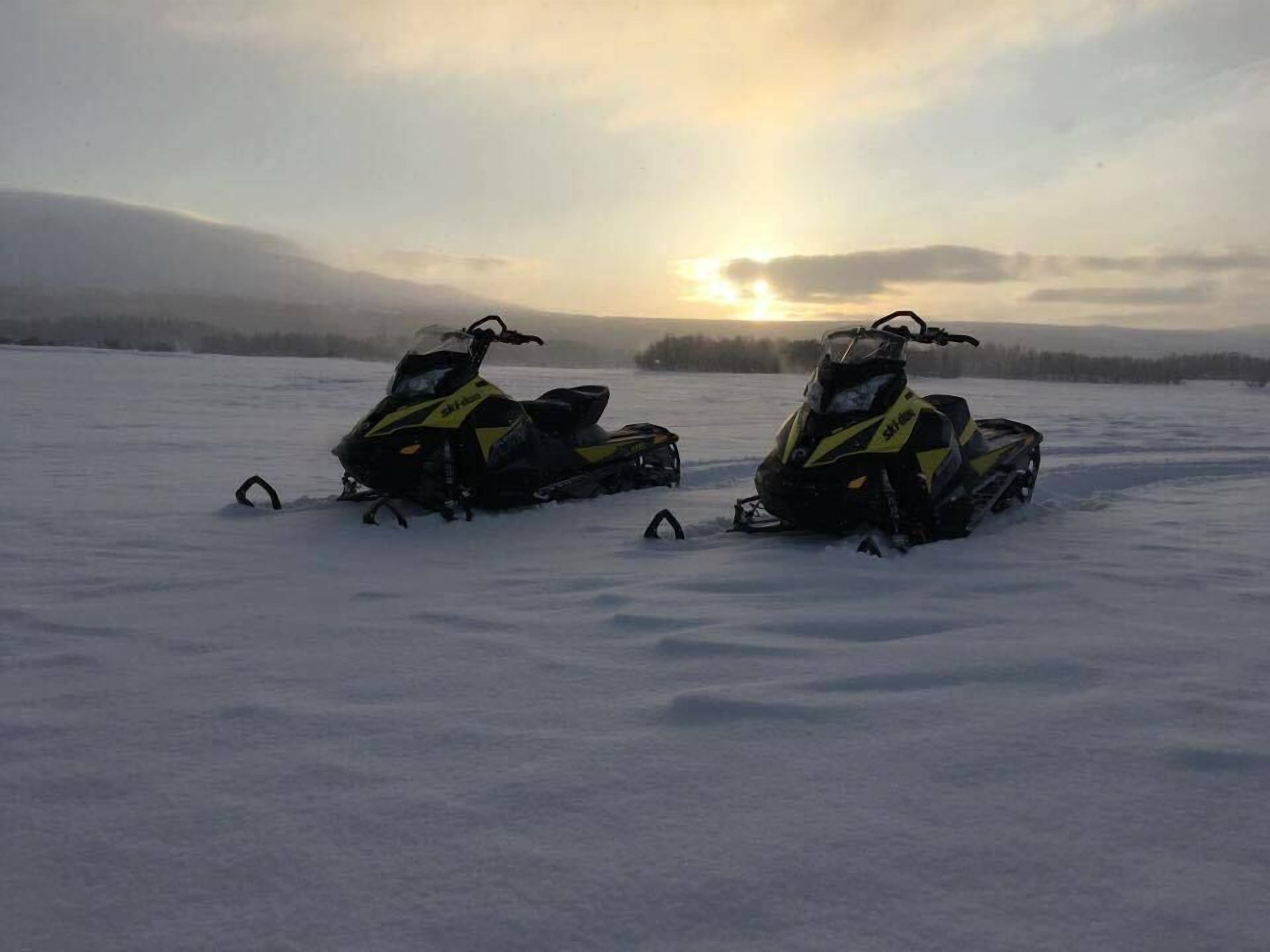 Två snöskotrar står parkerade på ett snötäckt fält med en solnedgång i bakgrunden.