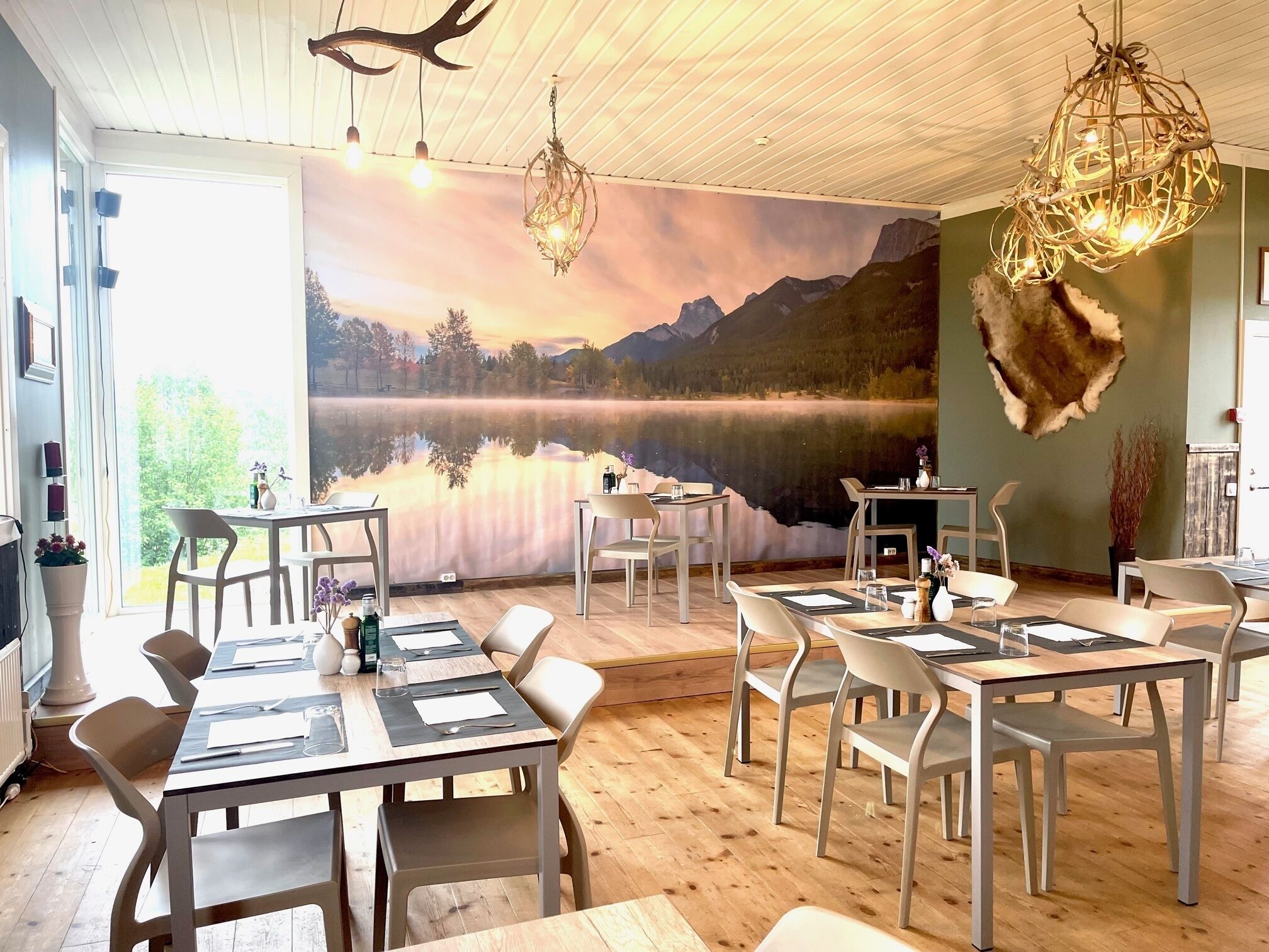 Zona pranzo con tavoli e sedie, un affresco raffigurante un lago, e un soffitto con travi in legno e lampade a sospensione.