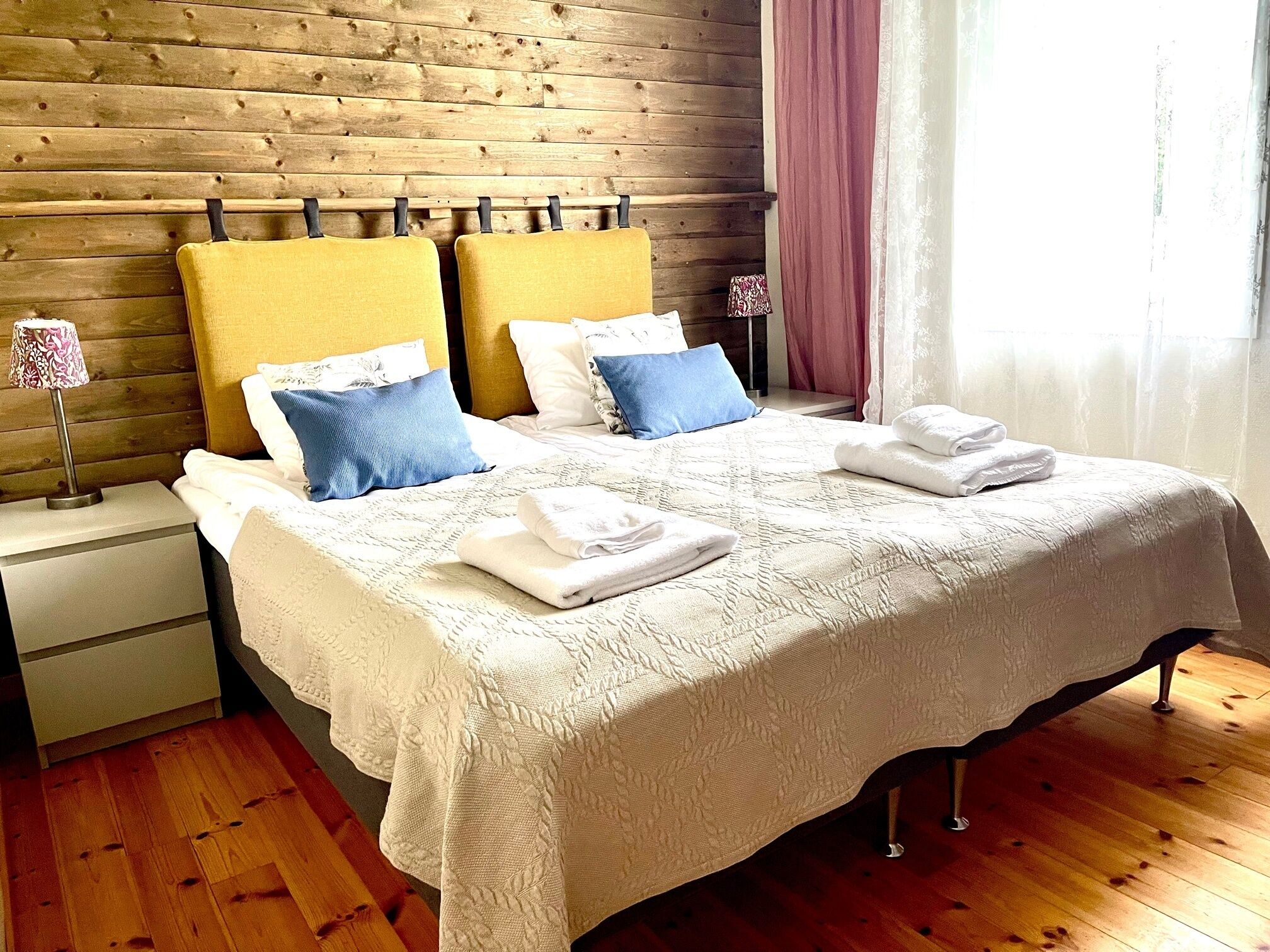 A double bed with a white quilt, two yellow headboards, and blue pillows. There are two bedside tables with lamps, a wooden floor, and a window with curtains.