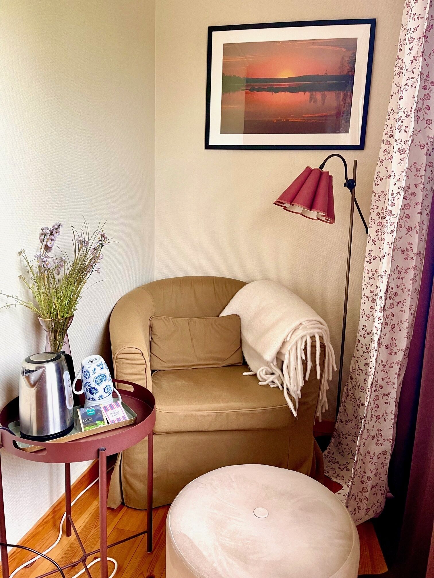 Een gezellige kamer met een beige bank, een klein rond voetenbankje, een tafel met een waterkoker, een mok en een vaas bloemen. Er hangt een ingelijst beeld aan de muur en staat een vloerlamp.
