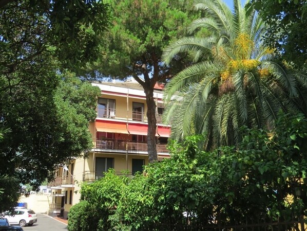 Un edificio de varios pisos con balcones, rodeado de abundante vegetación, incluyendo una palmera.