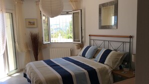 A bedroom with a bed, a bedside table, a nightstand, a window with curtains, a radiator, a wall-mounted mirror, and a view of greenery outside.