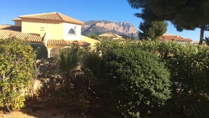 A yellow building with a tiled roof, surrounded by greenery and mountains in the background.