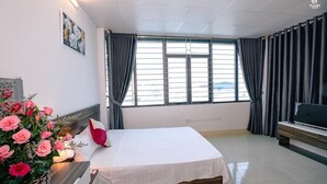 A hotel room with a bed, a TV, a window with curtains, a wall-mounted picture, and a floral arrangement. - KorHan Apartment (Hanoi)