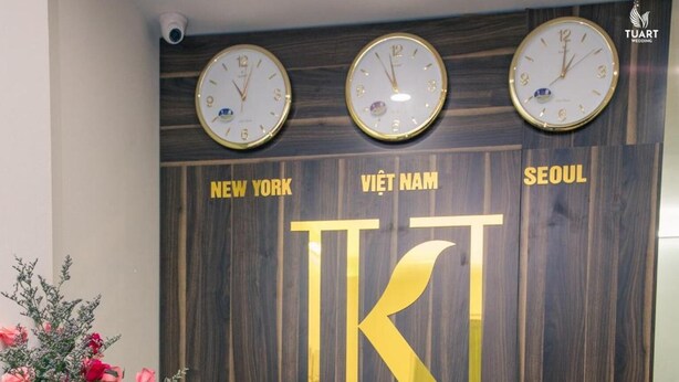 A hotel reception desk with a wooden backdrop displaying the name 'KI' and 'KORHAN HOTEL APARTMENT'. There are clocks showing different times, and a potted plant with pink flowers. - KorHan Apartment (Hanoi)