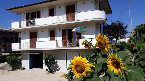 A two-story building with a balcony, a door, and a small garden with sunflowers.