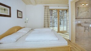 Habitació amb un llit de fusta, un bany amb un lavabo i mirall, i vista cap a l'exterior a través d'una porta de vidre.