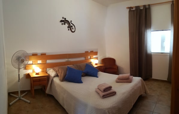 Un dormitorio ordenado con cama, lámparas de noche, ventilador de pared y una ventana con cortinas.
