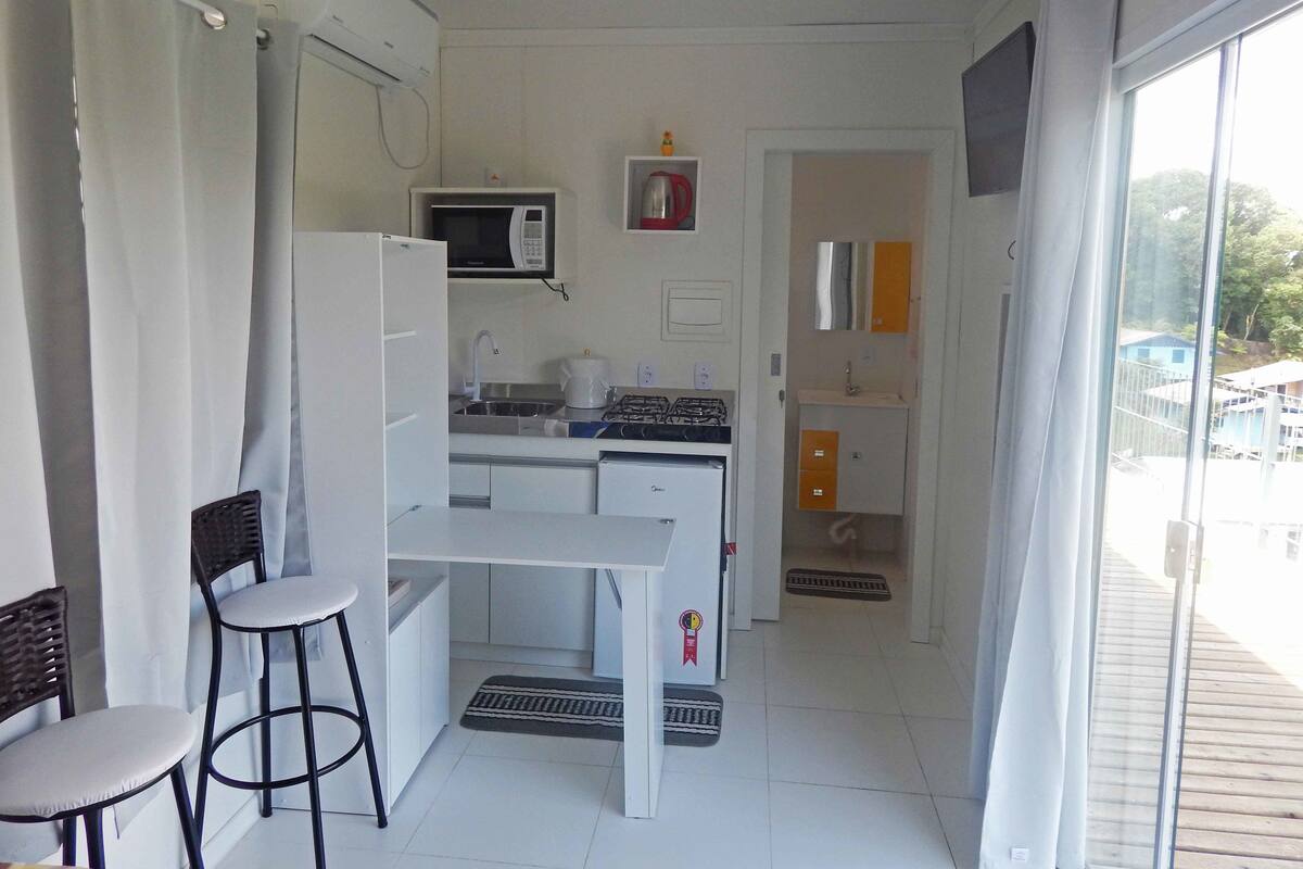 Une cuisine compacte équipée d’un four à micro-ondes, d’un évier et d’une cuisinière à gaz. On y trouve deux tabourets de bar et un petit réfrigérateur. Une fenêtre offre une vue sur l’extérieur.