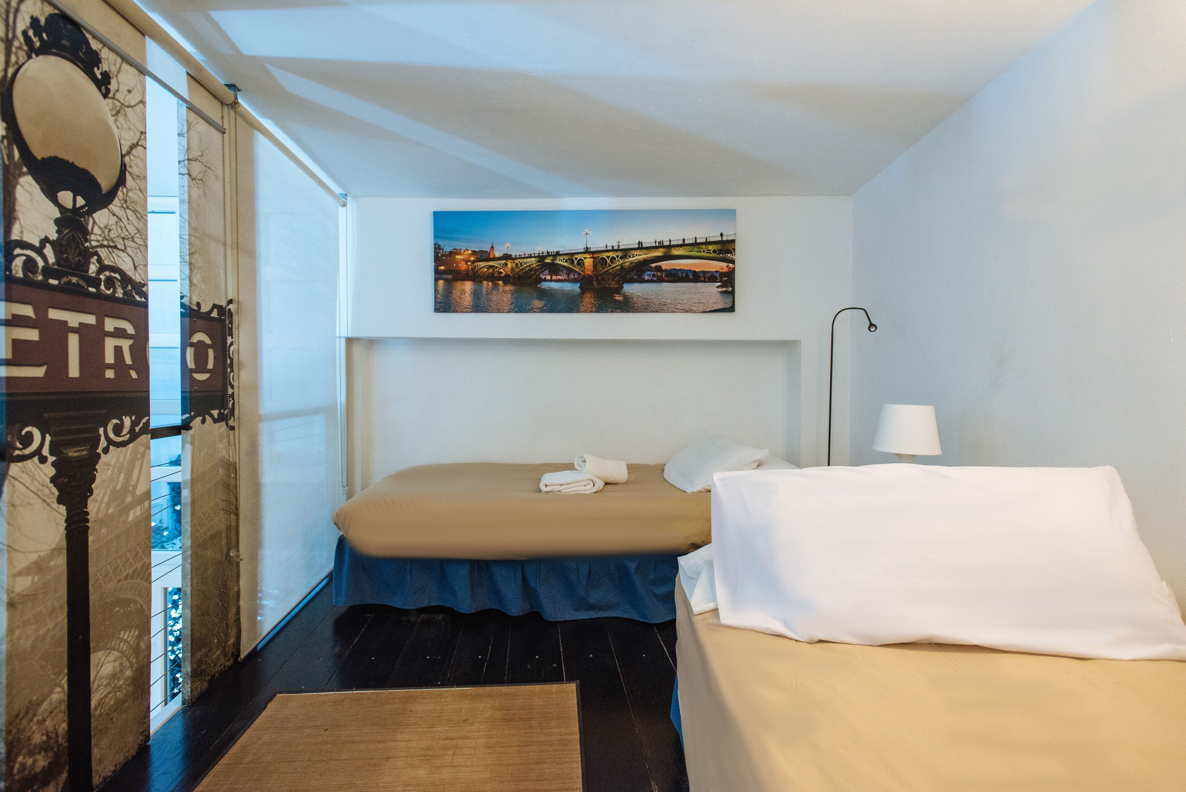 A bedroom with a bed, a nightstand, a lamp, a painting of a bridge, and a window with curtains.