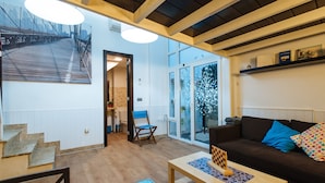 A modern living room with a wooden staircase, a brown sofa with blue cushions, a glass coffee table, and a large window with a view of a bridge.