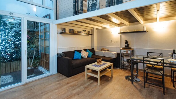 A modern living space with a glass door leading to a garden, a dark-colored sofa with colorful cushions, a wooden coffee table, and a dining area with a glass table and metal chairs.