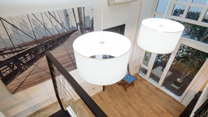 A high-ceilinged room with a large window overlooking a cityscape, a wooden floor, and a staircase with a black railing.