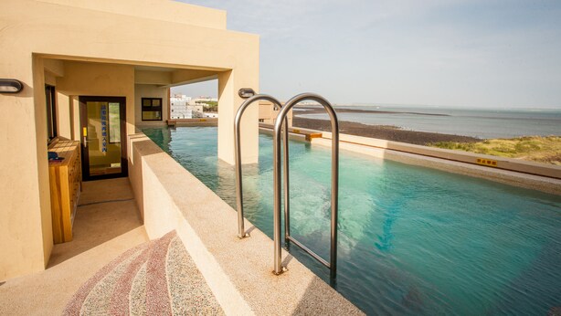 Una piscina con barandillas de metal, una puerta que da a un edificio y vistas a la playa.