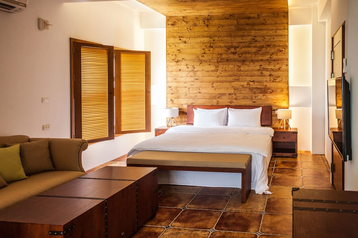 Un dormitorio con techo de madera, una cama con ropa de cama blanca, un banco de madera y una mesita de noche con una lámpara.