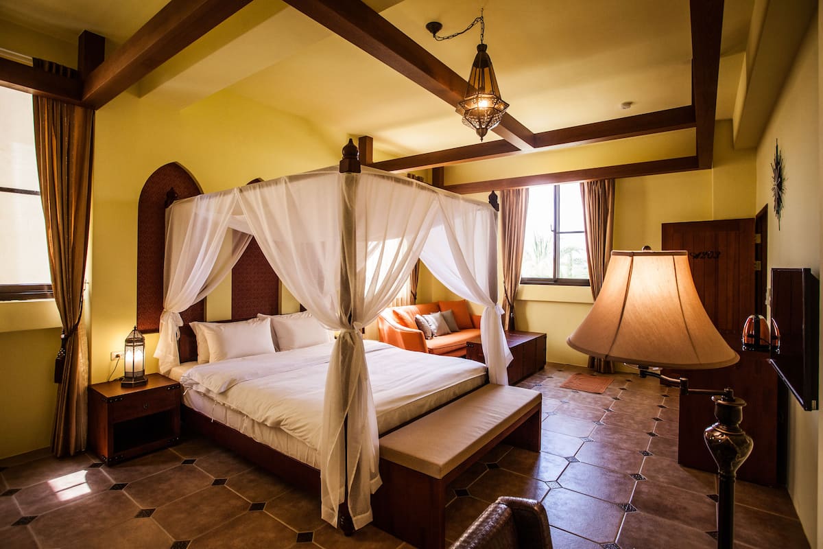 Una cama con dosel y cuatro columnas, con una mesita de noche, una lámpara y vistas al exterior a través de una ventana.