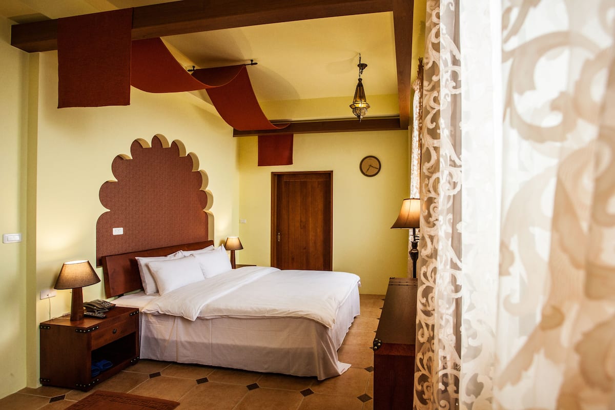 Un dormitorio con una cama grande, cabecera de madera, dos mesitas de noche con lámparas, una puerta de madera y un reloj en la pared.