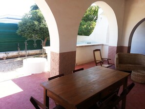 Un patio con mesa y sillas de madera, vistas a zonas verdes y una masa de agua, además de una pared blanca con una ventana.