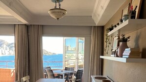 A dining area with a table and chairs, a kitchen with white cabinets, and a view of the sea and buildings.