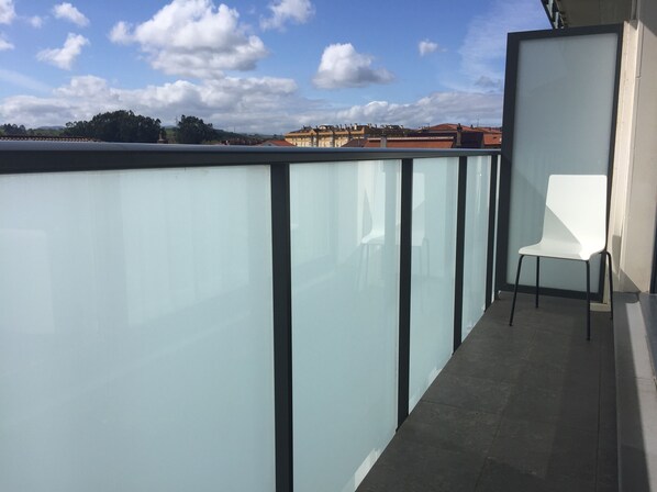 A balcony with a glass partition, a white chair, and a view of buildings in the distance.