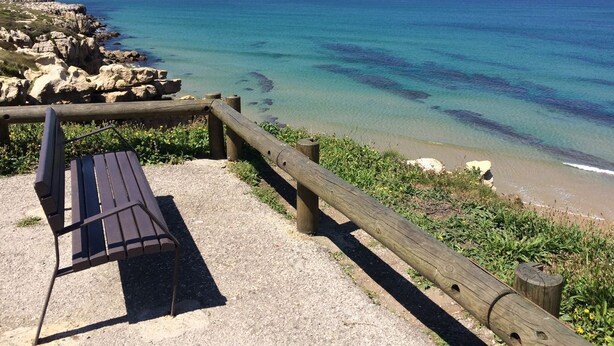 Un banco de madera con vistas a una playa, agua azul cristalina y costa rocosa.
