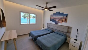 Un dormitorio con dos camas, un ventilador de techo, una ventana con vista al océano y un cuadro en la pared.