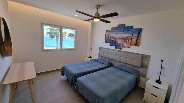 A bedroom with two beds, a ceiling fan, a window with a view of the ocean, and a painting on the wall.