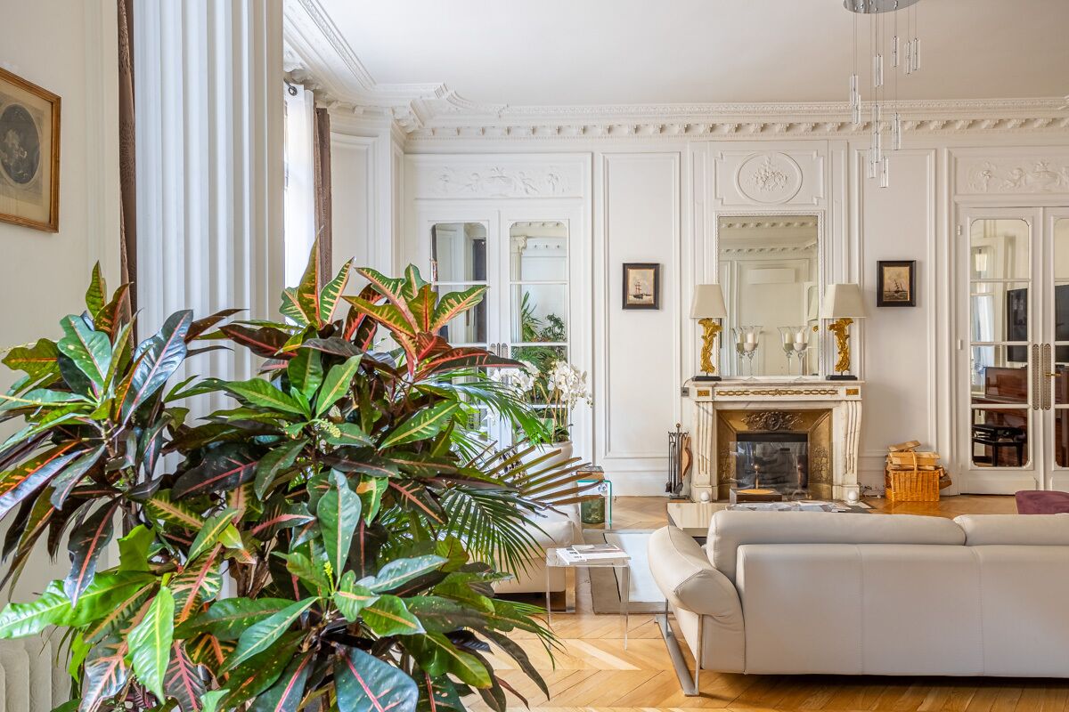 A spacious living room with a fireplace, white furniture, and large green plants.