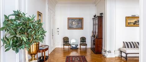 A room with a wooden floor, a large green plant, two chairs, a sofa, and a painting on the wall.