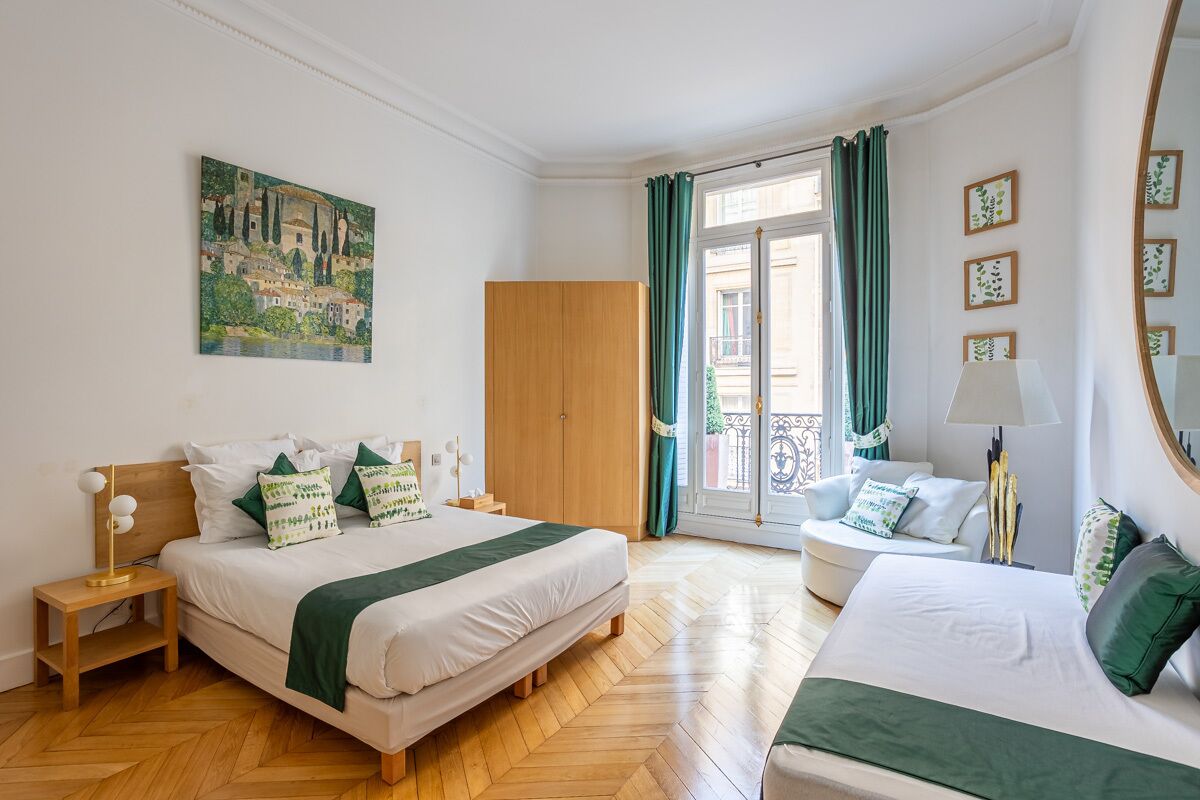 Habitación de hotel con cama, mesita de noche con lámpara, una silla, armario, un cuadro en la pared y balcón con cortinas.