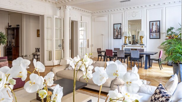 Ein geräumiges Wohnzimmer mit einer Couch, einem Couchtisch und Orchideen. Es gibt eine Essecke mit Stühlen und einem Tisch, und im Hintergrund eine Küche.