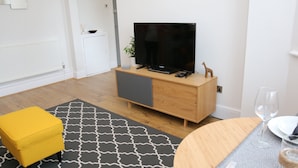 Una sala de estar moderna con un aparador de madera, un televisor de pantalla plana, un reposapiés amarillo y una mesa de comedor con un vaso de agua.