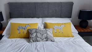 Una cama con ropa de cama blanca, dos almohadas amarillas con diseño de cebra, una almohada gris estampada y dos lámparas negras sobre mesitas de noche de madera.
